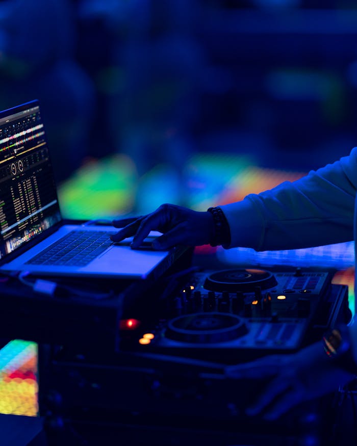 DJ using a mixer and laptop at a vibrant nightclub with blue lighting.